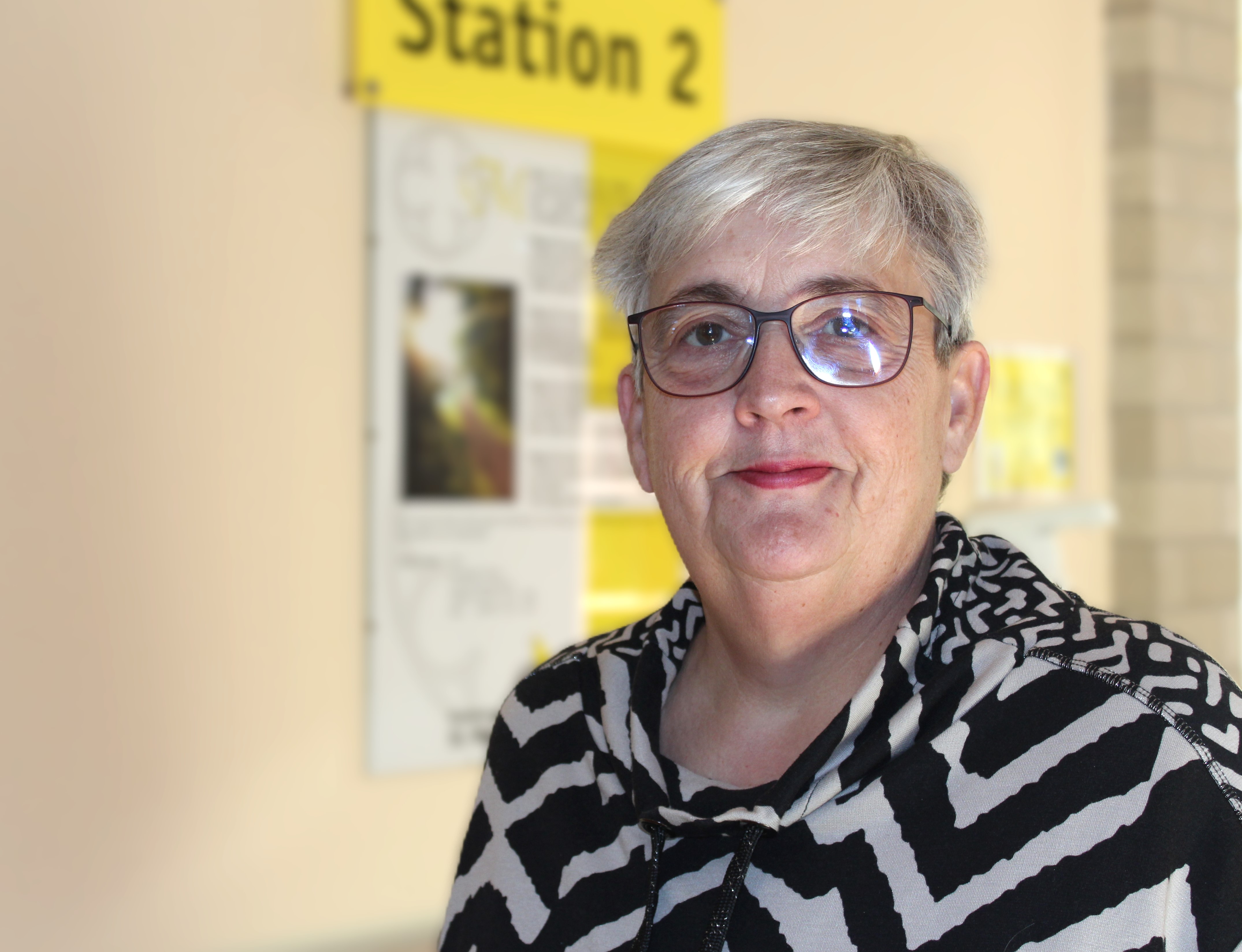 Die Patientenfürsprecherin Silvia Roßbach steht freundlich lächelnd und zugewandt vor einem gelben Stationshinweisschild. 