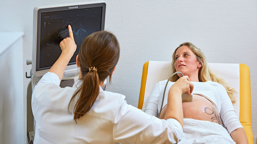 Eine Gyäkologin macht eine Ultraschalluntersuchung auf dem Bauch einer Schwangeren. Sie zeigt auf den Bildschirm.