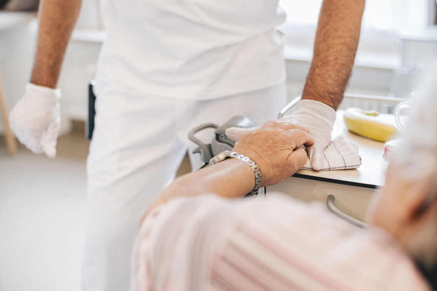Ein Patient, im Bett liegend, legt dankend seine Hand auf die eines Mitarbeiters der Reinigung, der gerade ein Fläche reinigt. Die Gesichter sind nicht erkennbar.
