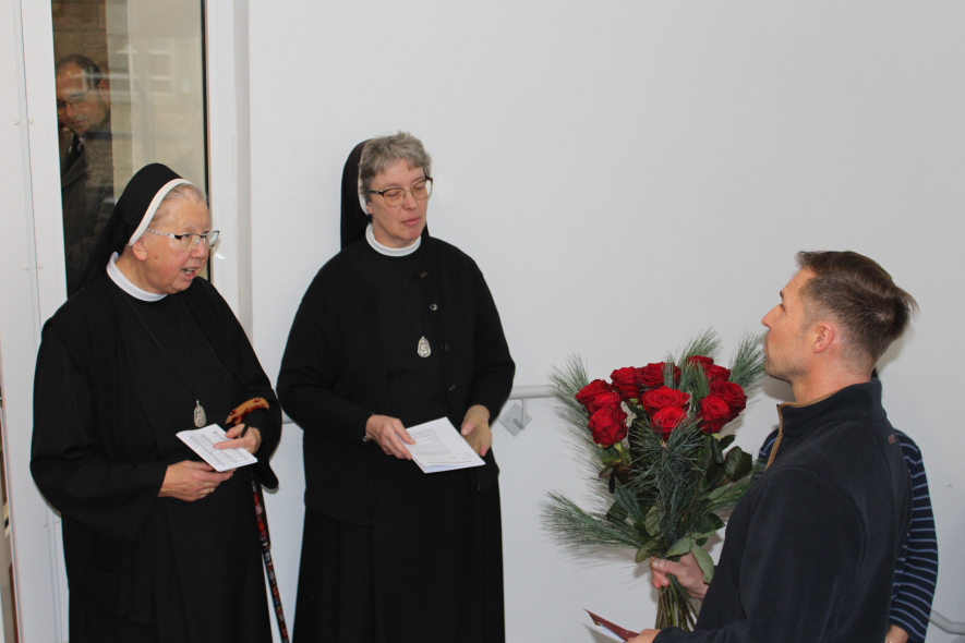 Der Wertebeiratsvorsitzende Markus Neuberg überreicht lächelnd und zugewandt einen großen Strauß roter Rosen an die Ordensfrau Schwester Dominika und Schwester Teresa. Beide sind sichtlich ergriffen.