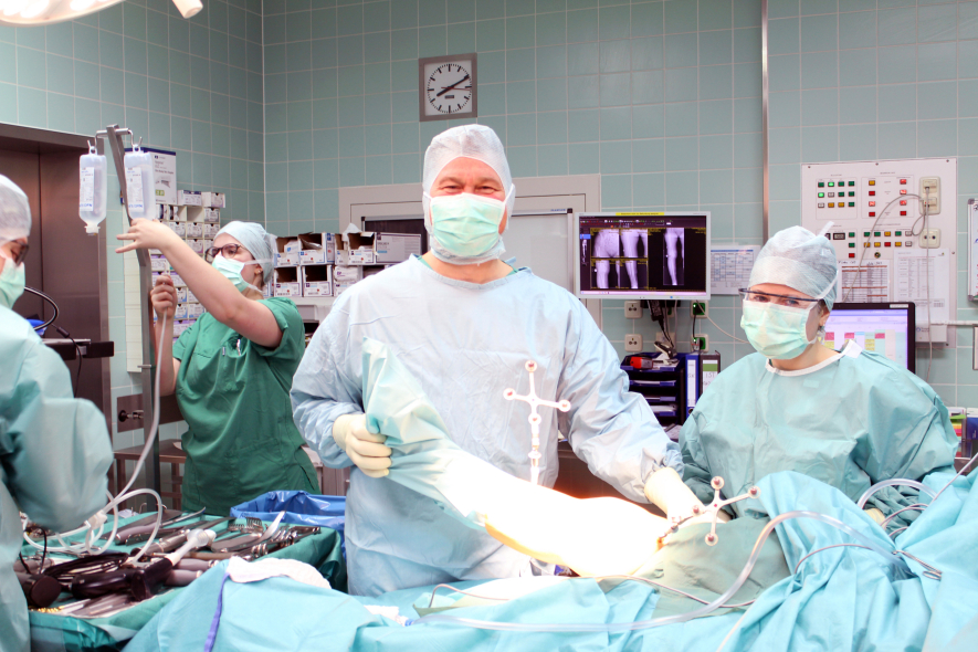 Im Orthopädie-OP-Saal schaut Herr Dr. John, der einen Mundschutz und OP-Kleidung trägt, in die Kamera. Um ihn herum stehen seine Kollegen.