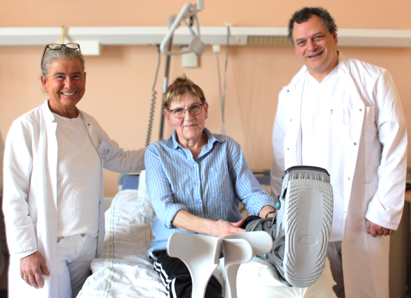 Frau Dr. Schenk, Carola Poppe, die eine Beinorthese trägt und im Krankenbett liegt und Dr. Lietz lächeln in die Kamera.