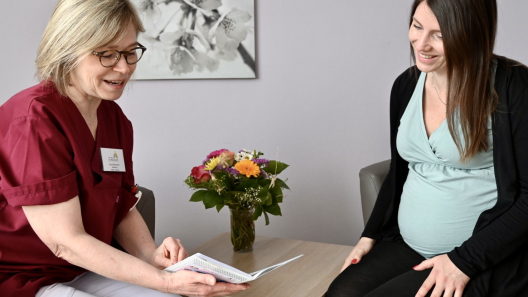 Hebamme Ulrike befindet sich im Bild links im Gespräch mit einer Schwangeren. Sie erklärt ihr freundlich und zugewandt die Aufnahmeformalitäten. Auf einem Beistelltisch steht ein bunter Blumenstrauß.ß auf dem 