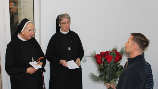Der Wertebeiratsvorsitzende Markus Neuberg überreicht lächelnd und zugewandt einen großen Strauß roter Rosen an die Ordensfrau Schwester Dominika und Schwester Teresa. Beide sind sichtlich ergriffen.