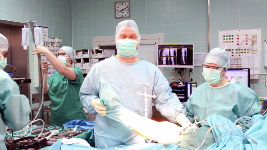 Im Orthopädie-OP-Saal schaut Herr Dr. John, der einen Mundschutz und OP-Kleidung trägt, in die Kamera. Um ihn herum stehen seine Kollegen.