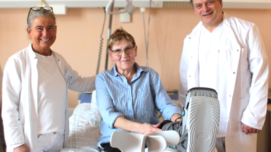 Frau Dr. Schenk, Carola Poppe, die eine Beinorthese trägt und im Krankenbett liegt und Dr. Lietz lächeln in die Kamera.